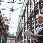Man in a suit holding documents in a vehicle repair facility, overseeing structural repairs with advanced technology and equipment.