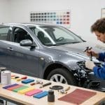 Auto body technician refinishing a gray car using spray paint, surrounded by color samples and tools, in a professional auto paint shop setting.