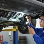 Mechanic inspecting vehicle undercarriage with flashlight, highlighting frame damage assessment in auto repair context.